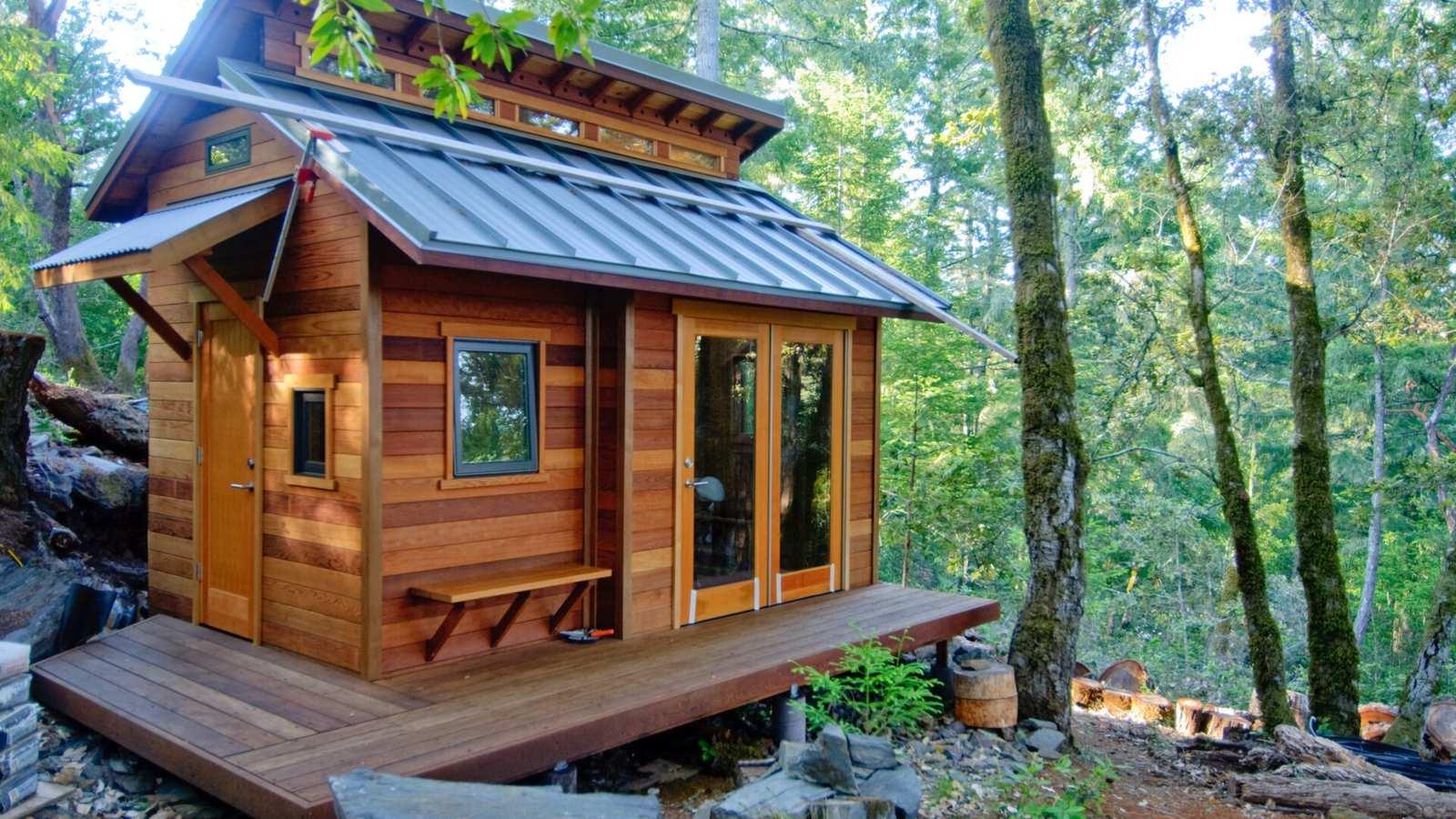 Petite maison en bois en forêt, avec une terrasse, une double porte vitrée et une partie haute éclairée par une rangée de fenêtres sous le toit